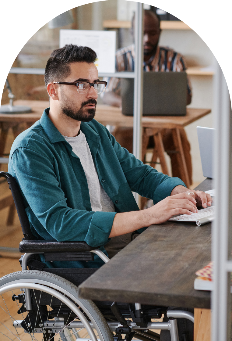 Accessibility training for employers. A man with short black hair and glasses sits in a wheelchair at a desk in his workplace and is using a keyboard. In the blurred background, another man can be seen working on a laptop as well.