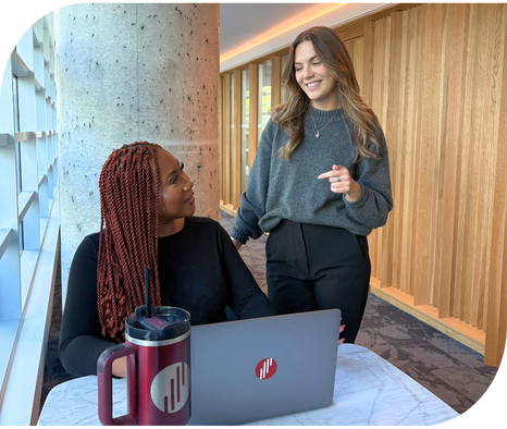 Secure your limited-time recruitment offer with Envol. Two women are smiling at each other in a professional workplace setting. The woman who is standing has long blonde hair, a grey sweater, and black pants. Beside her, another woman sits at a table working on a laptop. The woman sitting has long red braided hair and a black long sleeve shirt.